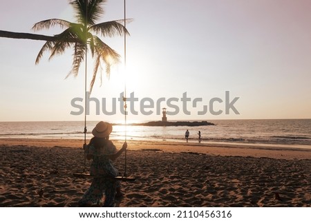 Woman traveller is siting on a swing and enjoy looking beautiful sunset with backgound of Nang Thong Beach in Khao lak lighthouse at Phang Nga Province, Thailand. 