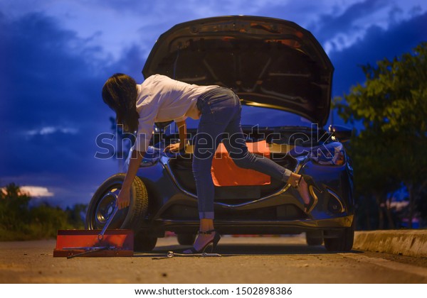 Woman try to fix problem of car by herself\
with belonging tools, need help and assistant at dark of the night,\
scary and worry alone in the dark, car engine failure or tire need\
replacement