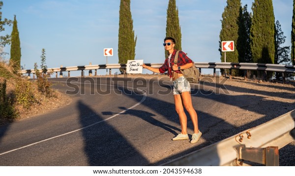 Woman try to stop car with\
cardboard sign, copy space. Girl with cheerful face travelling by\
hitchhiking with road on background. Travelling and hitchhiking\
concept.