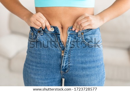 Woman Trying To Zip Up Too-Tight Jeans After Gaining Weight Standing At Home. Dieting And Weight-Loss Concept. Cropped