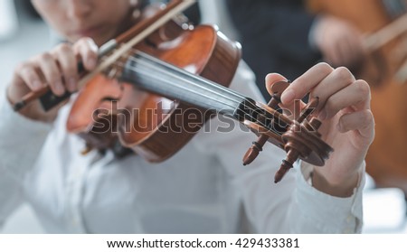 Woman tuning her violing and rotating pegs, cello player on background, selective focus