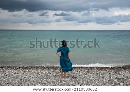 A woman in a turquoise dress stands alone on the pebble beach by the water and looks at the sea on a cloudy summer day