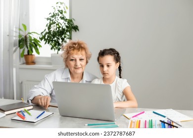 Woman tutor or foster parent mum helping cute caucasian school child girl doing homework sitting at table. Diverse nanny and kid learning writing in notebook studying at home. - Powered by Shutterstock