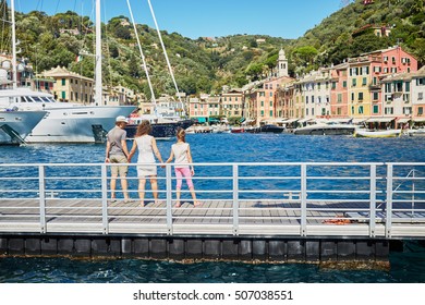 Woman and two children stand holding hands and looking at boats in sea bay with colorful buildings on the shore.