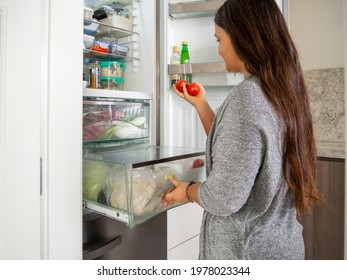 Woman With Two Tomatoes With Open Drawer In The Fridge. Healthy Eating. Food Container And Leftovers Inside A Domestic Refrigerator. Female Preparing A Salad And Looking For Ingredients At Home.