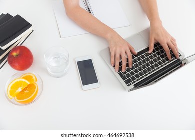 Woman typing at her laptop keyboard. Smart phone, glass of water, orange and apple lie on table beside her. Concept of hard work at corporation. Top view. Close up - Powered by Shutterstock