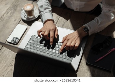 Woman typing on laptop in a café, with smartphone, notebook, sunglasses, and coffee on wooden table. Stylish and productive workspace in natural light.