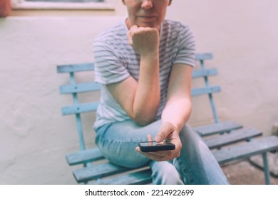 Woman Typing Text Message On Mobile Smart Phone While Sitting On Wooden Bench. Female Person Wearing Jeans Trousers And Stripped T-shirt Using Smartphone For Communication. Selective Focus.