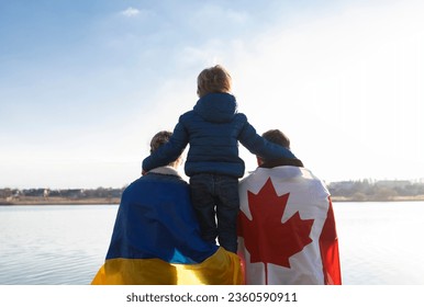 woman with Ukrainian flag, man with Canadian flag behind back and child , sitting with backs together in nature . International support and assistance to Ukraine. Problems of refugees and immigrants - Shutterstock ID 2360590911