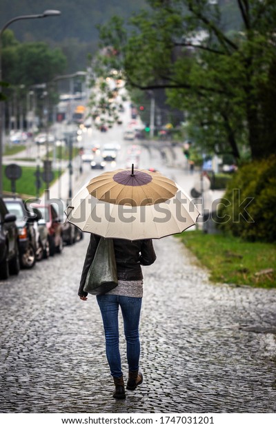 Woman\
with umbrella walking on road in city during rain. Trendy woman\
wearing street style casual clothing. City\
life