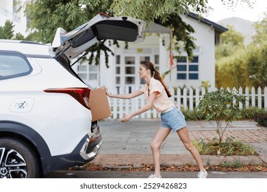 Woman unloading a cardboard box from the trunk of a white SUV car against a residential backdrop with greenery Sunny day, casual summer outfit, focused on her task - Powered by Shutterstock