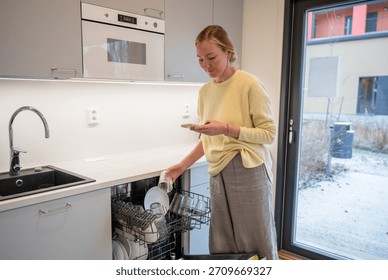 Woman unloading dishwasher while using smartphone in modern kitchen