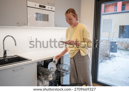 Woman unloading dishwasher while using smartphone in modern kitchen