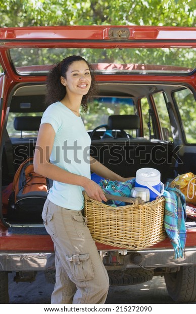 Woman unloading parked SUV on\
family camping trip, carrying picnic hamper, smiling,\
portrait