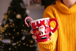 Woman Unrecognizable In Orange Sweater Hold Santa Claus Mug With Sugar Cane, Christmas Tree Background. 