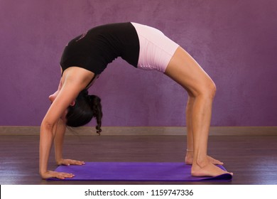 Woman In Urdhva Dhanurasana Yoga Asana. Female Yogi In Wheel Pose On Purple Mat Indoors. Young Lady In Upward Bow Posture. Advanced Practitioner, Backbend, Flexibility Concepts