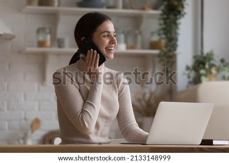 Woman use cellphone enjoy communication sit in kitchen at table with laptop, smile looks aside, ordering food, goods remotely, make call by work, having personal conversation. Tech, connection concept