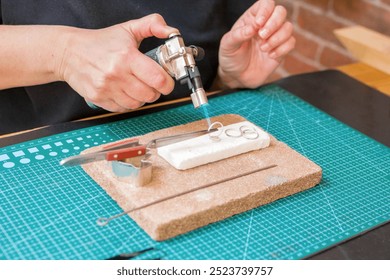 A woman uses a blowtorch to create jewelry on a on a worktable with cutting mat. - Powered by Shutterstock