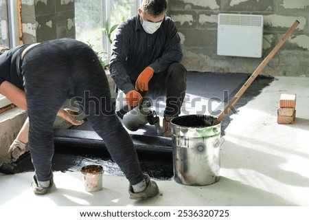 A woman uses a brush to apply bitumen to a concrete floor, waterproofing a floor in a house.