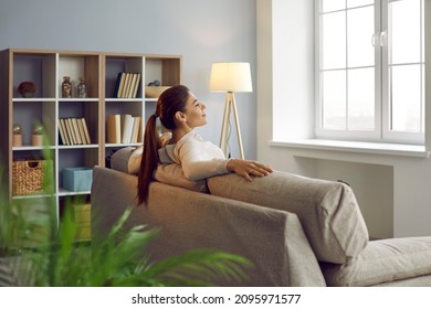 Woman uses chance to relax, unwind and get some peace of mind on comfy couch at home. Happy lady enjoying silence and quiet leisure time in beautiful room with comfortable sofa and Nordic wooden shelf