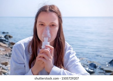 Woman Uses Nebulizer On Seaside. Inhaling Through Inhaler Mask. Fibrosis Cystic Copd And Treatment Inhaler. Asthma Pulmonary, Respiratory Breath Problem And Cure. Painkiller Sedative Gas Oxygen.