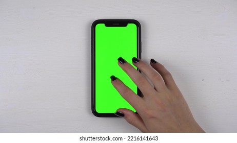 A Woman Uses A Smartphone Lying On The Table. Green Screen On Phone. The Girl Taps Her Finger On The Display. Hand Close Up.