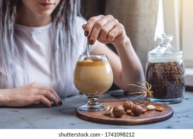 A woman uses a spoon to stir the whipped mousse from Dalgona coffee in a clear cup. Delicious cold drink. Trendy spectacular drink that can be prepared at home