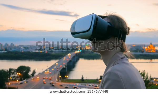 Woman uses virtual reality glasses in the city\
after sunset. Evening time, twilight. Relax, entertainment and\
technology concept
