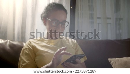 Woman using app on mobile phone at home. Beautiful relaxing girl relaxing while chatting on the cell phone. Young caucasian woman in the sun using a smartphone while sitting on the sofa in rays of sun