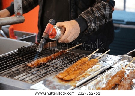 Woman Using a Blow Torch on a Very Long Skewer of Grilled Sweet and Spicy Chicken at a Street Food Stall in South Korea