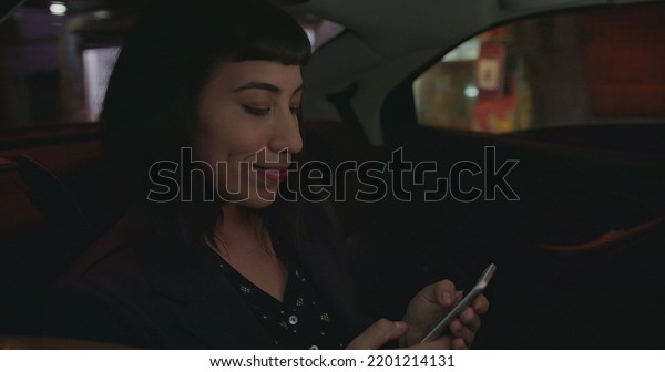 Woman using cellphone in car backseat at night.\
Person riding taxi cab in urban city street typing message on\
smartphone device commuting from\
work
