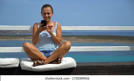 Woman using cellphone texting sms message outside on balcony terrace of luxury hotel by the sea
