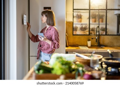 Woman using digital tablet hanging on the wall in the kitchen, controlling smart home devices. Modern kitchen interior with food in front - Powered by Shutterstock