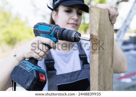 A woman is using an electric drill on a piece of wood at construction site, implying maintenance or setup