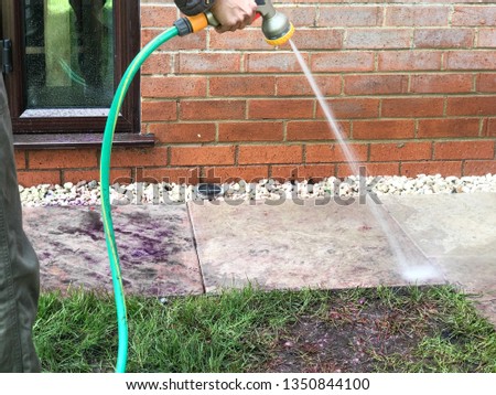 A woman using a garden hose in the garden at home, on a sunny day, washing away colours and mess to clean the garden. 