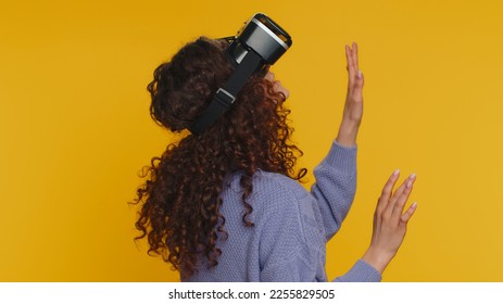 Woman using headset helmet to play simulation game app online. Watching virtual reality 3D 360 video content. Girl in VR goggles isolated on studio yellow background. Futuristic technology. Rear view - Powered by Shutterstock