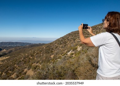 A Woman Using Her Cell Phone To Take A Picture Of The Valley Below After A Difficult Hike