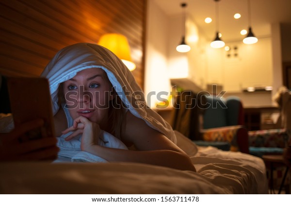 Woman using her phone under blanket in bed at\
night. Woman using her phone in bed at night. Light of the screen\
illuminating on her face. Technology, internet, communication and\
people concept