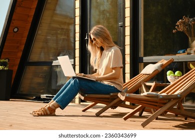 Woman using laptop sitting in a folding wooden chair on the terrace of wooden cabin - Powered by Shutterstock