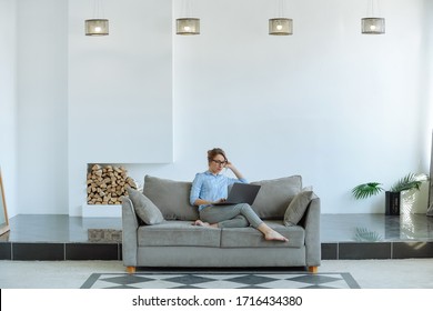 Woman using laptop sitting on a couch in a big bright minimalistic living room. Self-isolation.
