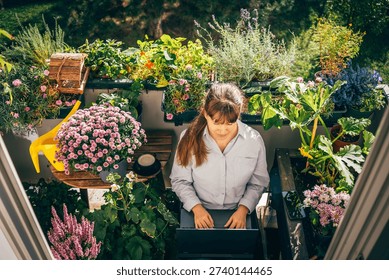 Woman using a laptop while sitting on a chair surrounded by blooming flowers and green plants on a balcony. Natural sunlight, cozy atmosphere. Concept of freelance work, work from home