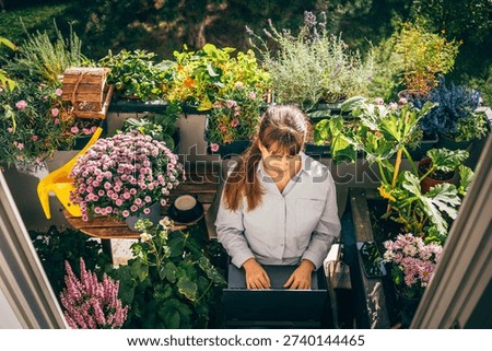 Woman using a laptop while sitting on a chair surrounded by blooming flowers and green plants on a balcony. Natural sunlight, cozy atmosphere. Concept of freelance work, work from home
