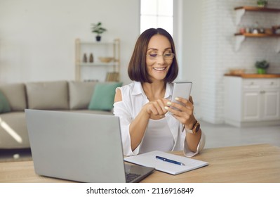 Woman using mobile phone. Happy beautiful lady in glasses sitting at desk with laptop, notebook and pen at home, holding cellphone, looking at screen, sending messages, or organizing business schedule