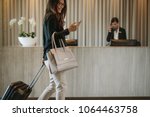 Woman using mobile phone and pulling her suitcase in a hotel lobby. Female business traveler walking in hotel hallway.