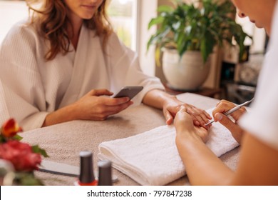 Woman Using Mobile Phone In Salon Receiving A Manicure And Nail Care Procedure By Beautician. Female Getting Nails Done By Manicurist At Beauty Salon.