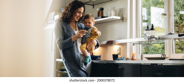 Woman Using Mobile Phone While Carrying Baby At Kitchen Counter. Mid Adult Mother With Newborn Boy Standing At Home.