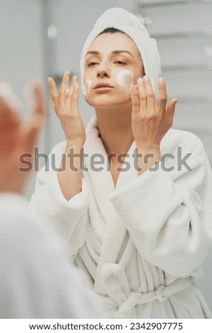 Woman using moisturizing foam for face washing, morning skincare routine. Adult female wearing towel on head looking in mirror in bathroom. Preparation for make up