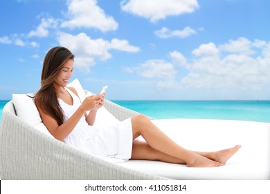 Woman Using Phone App Relaxing On Sun Bed On Outdoor Terrace. Home Living Outside Patio Furniture Asian Girl On Smartphone Lying On Beach Lounger In White Dress With Ocean Background Enjoying Summer. 