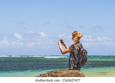 Woman Using Phone Camera On Beach With Smartphone Mobile Phone Against Teal Waters And Clouds. Beach Bag And Video Camera In Hand. Portrait