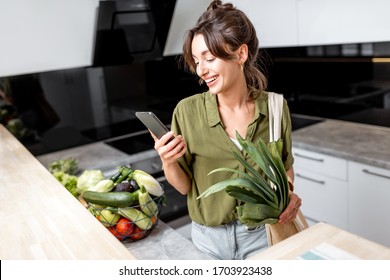 Woman Using Phone While Standing With Shopping Bag And Healthy Fresh Food On The Kitchen At Home